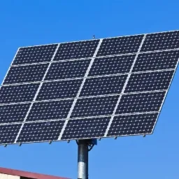 Panel fotowoltaiczny składający się z 20 ogniw, zamontowany na metalowym słupie, na tle błękitnego nieba. Widoczny fragment dachu budynku.