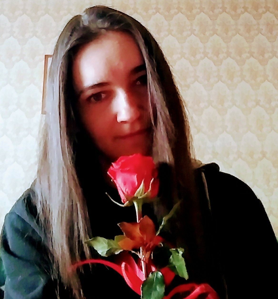 Kobieta z długimi włosami trzyma czerwoną różę na tle wzorzystej tapety. Ujęcie z bliska, skupiające się na twarzy i kwiatku.