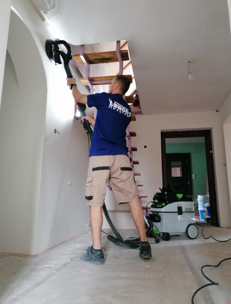 Pracownik szlifuje ścianę szlifierką z odkurzaczem, przygotowując ją do malowania. Widoczne schody i drzwi w tle. Fachowiec w trakcie pracy.