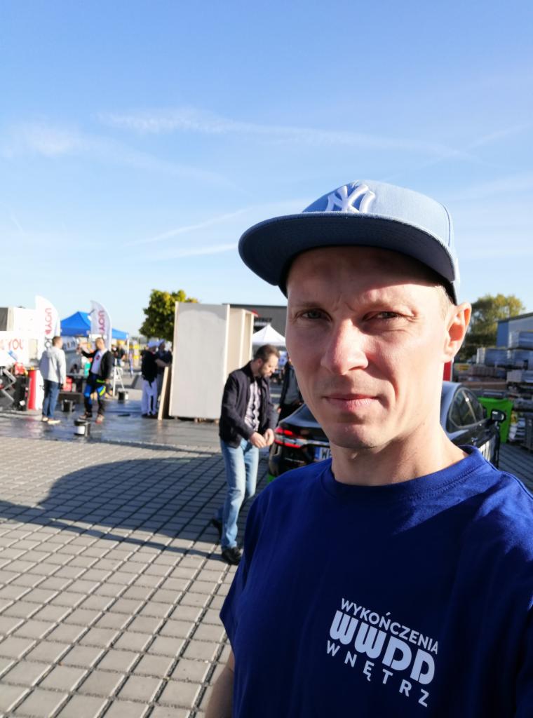 Mężczyzna w czapce NY i koszulce z logo 'Wykończenia Wnętrz WWDD' na tle budowy z płyt karton-gips, prawdopodobnie podczas dnia otwartego firmy.