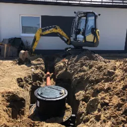 Żółta mini koparka stoi na skraju wykopu, w którym umieszczono czarny zbiornik z zieloną pokrywą i pomarańczową rurą, na tle białego domu z czarnym akcentem.