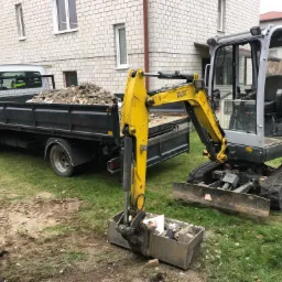 Żółto-szara mini koparka Wacker Neuson z łyżką wypełnioną gruzem, obok zaparkowany samochód ciężarowy z czarną skrzynią ładunkową, również wypełnioną gruzem, na trawiastym podłożu z widocznymi...