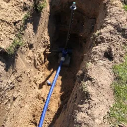 Głęboki wykop w piaszczystej ziemi z widoczną niebieską rurą wodną i pionową czarną rurą z zaworem na górze, element instalacji wodnej na betonowym fundamencie.