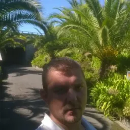 Mężczyzna na tle tropikalnej roślinności: palmy i krzewy w słoneczny dzień. W tle widoczna droga i tunel.