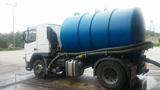 Biała ciężarówka z niebieskim zbiornikiem na płyny, zaparkowana na betonowym placu, widoczne węże i osprzęt.