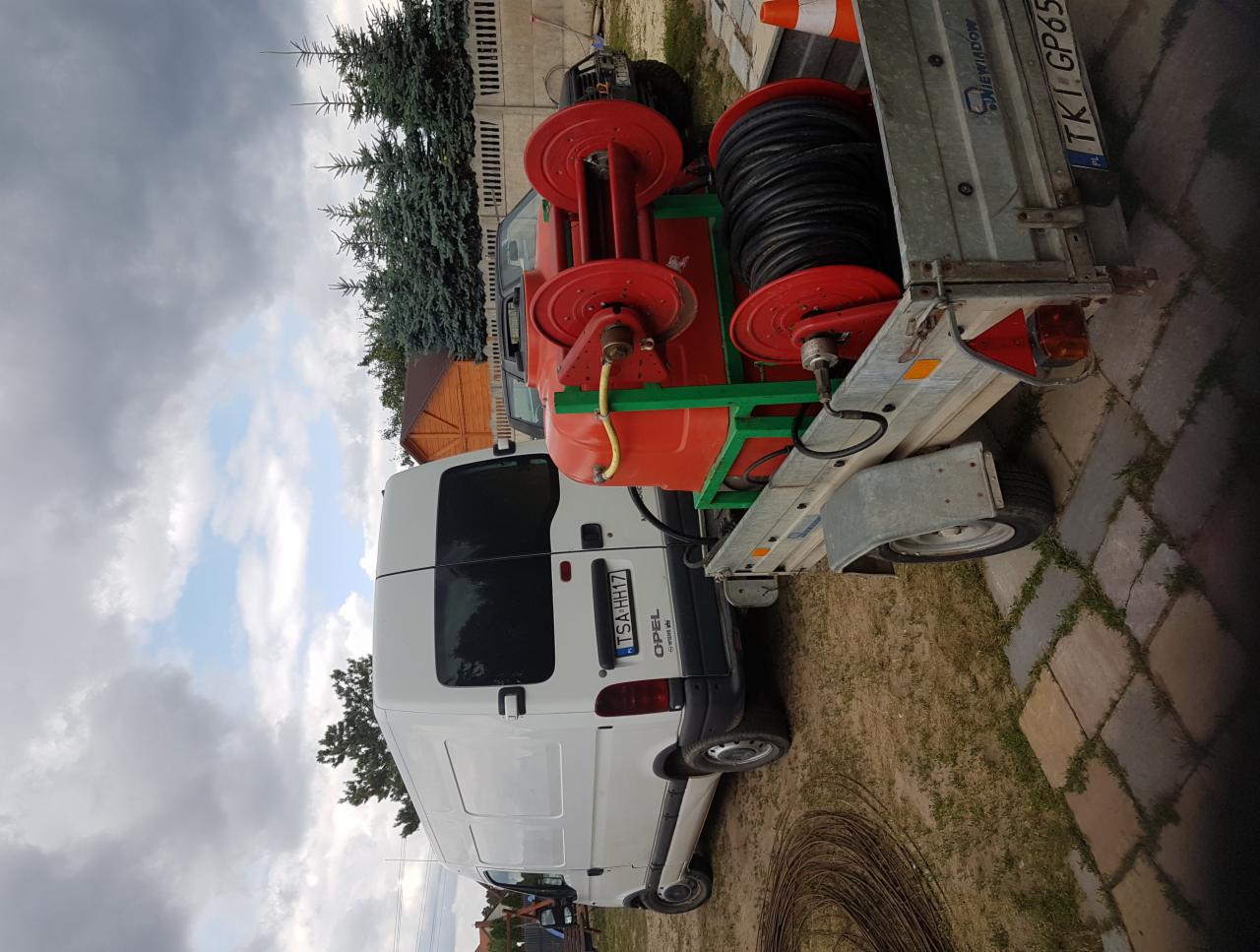 Biały furgon Opel z przyczepą zawierającą pomarańczowy bęben z nawiniętym czarnym kablem, zaparkowany na kostce brukowej obok trawnika i ogrodzenia.