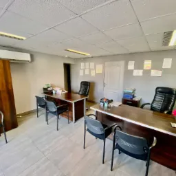 Biuro agencji nieruchomości z dwoma biurkami, krzesłami, kwiatami i dokumentami, oświetlone panelami LED.