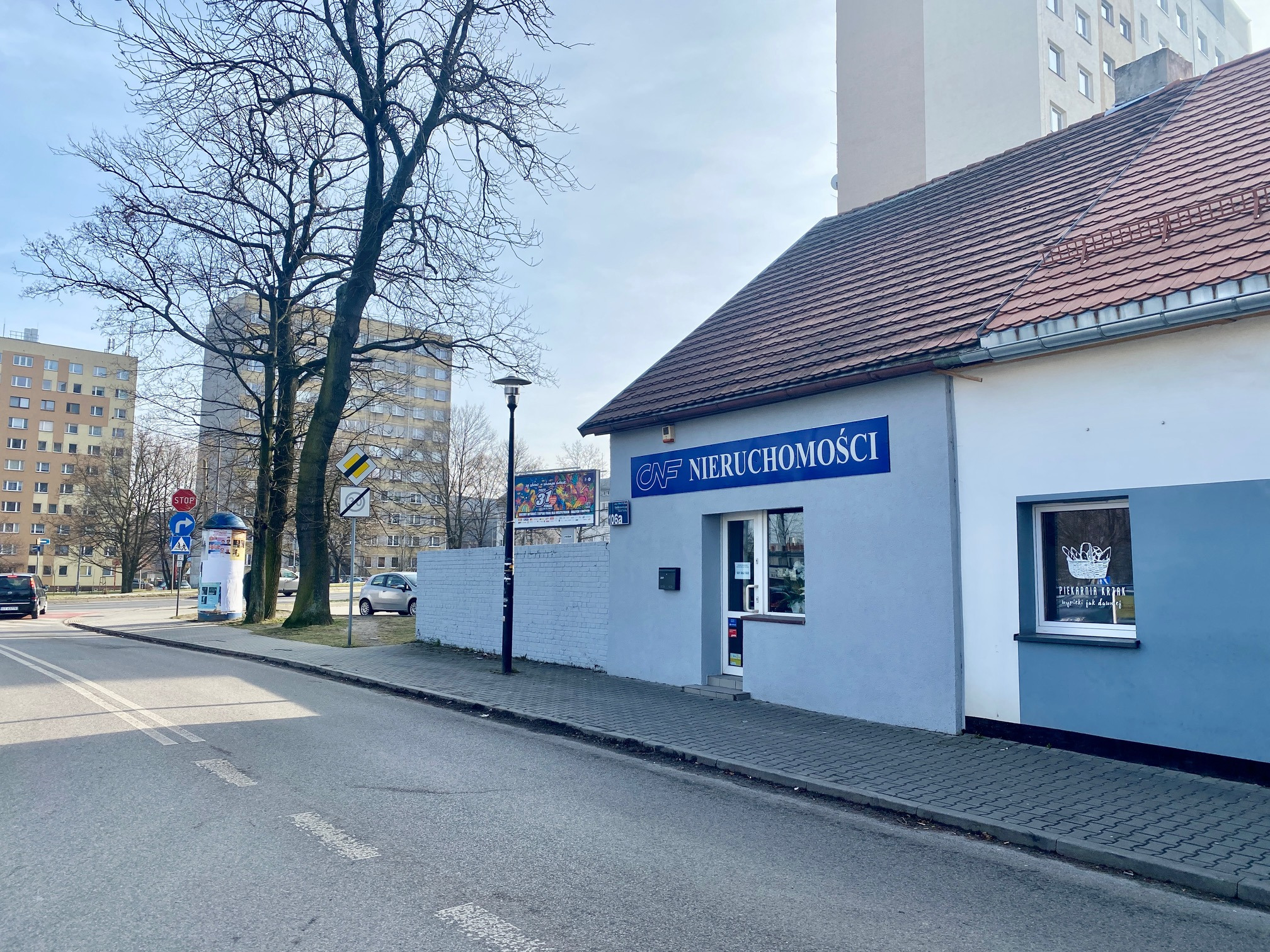 Budynek biura nieruchomości z szyldem 'CNF Nieruchomości' w jasnoniebieskim kolorze, widok z ulicy, w tle bloki mieszkalne i drzewa bez liści.