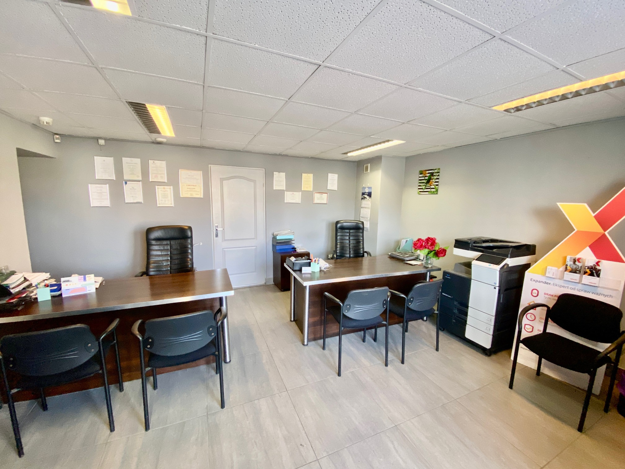 Wnętrze biura agencji nieruchomości z dwoma biurkami, krzesłami, drukarką i materiałami reklamowymi na stojaku; na ścianach wiszą certyfikaty.