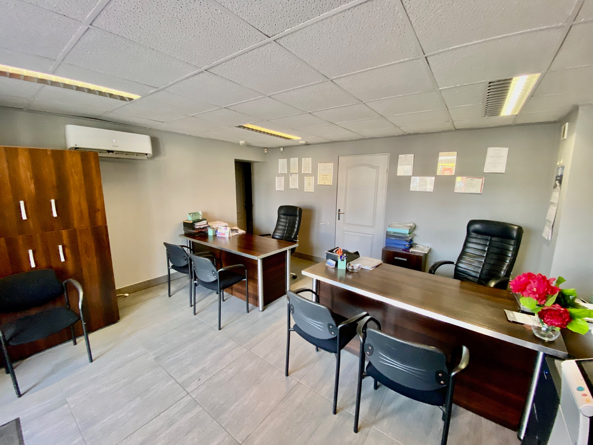 Biuro agencji nieruchomości z dwoma biurkami, krzesłami, kwiatami i dokumentami, oświetlone panelami LED.