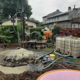 Wiercenie otworu w ziemi przy użyciu wiertnicy gąsienicowej; w tle widoczne budynki mieszkalne, zbiorniki na wodę i elementy ogrodu.