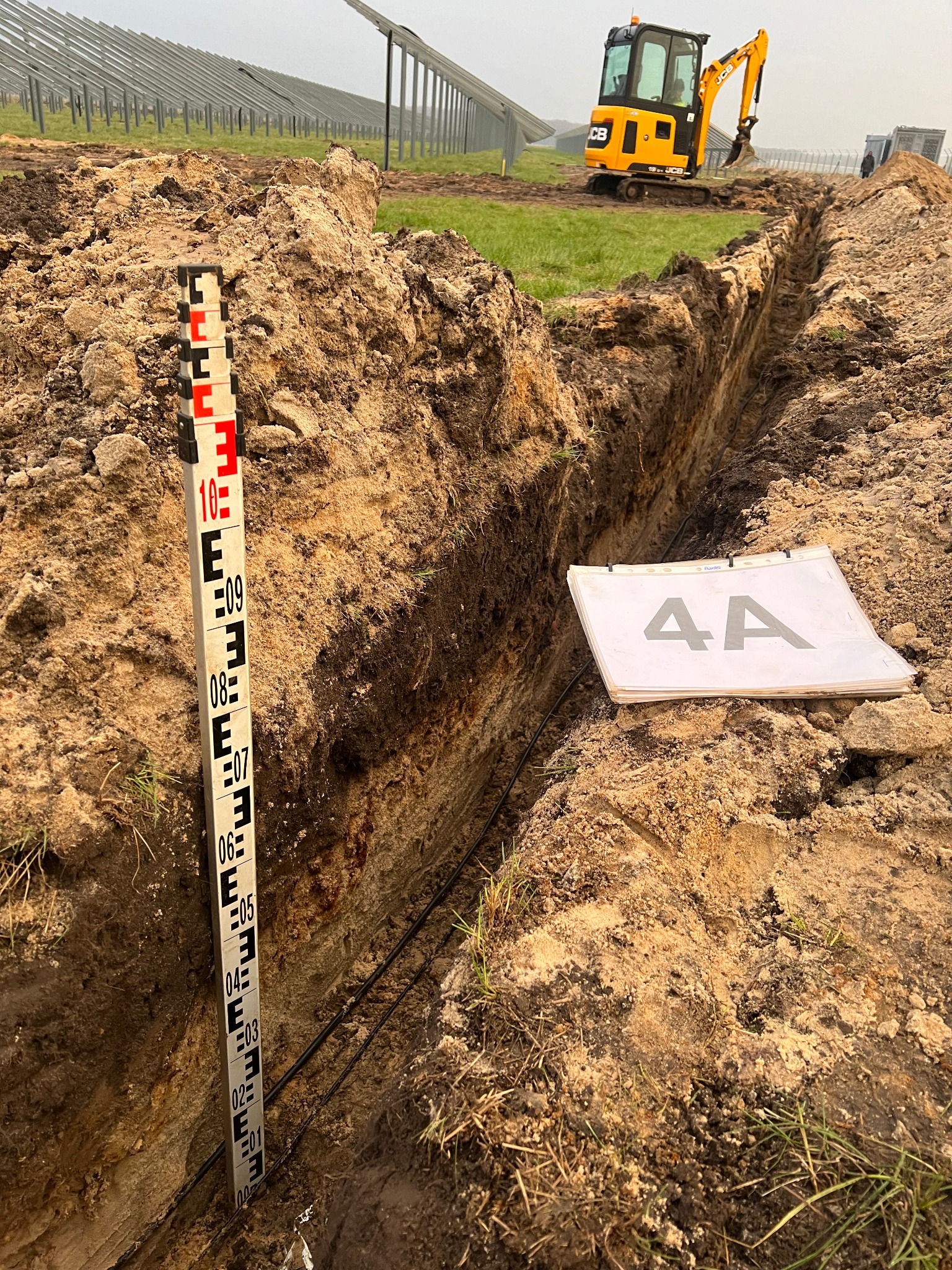 Wykopany rów z ułożonymi kablami, obok łata niwelacyjna z podziałką, w tle koparka i konstrukcja paneli fotowoltaicznych.