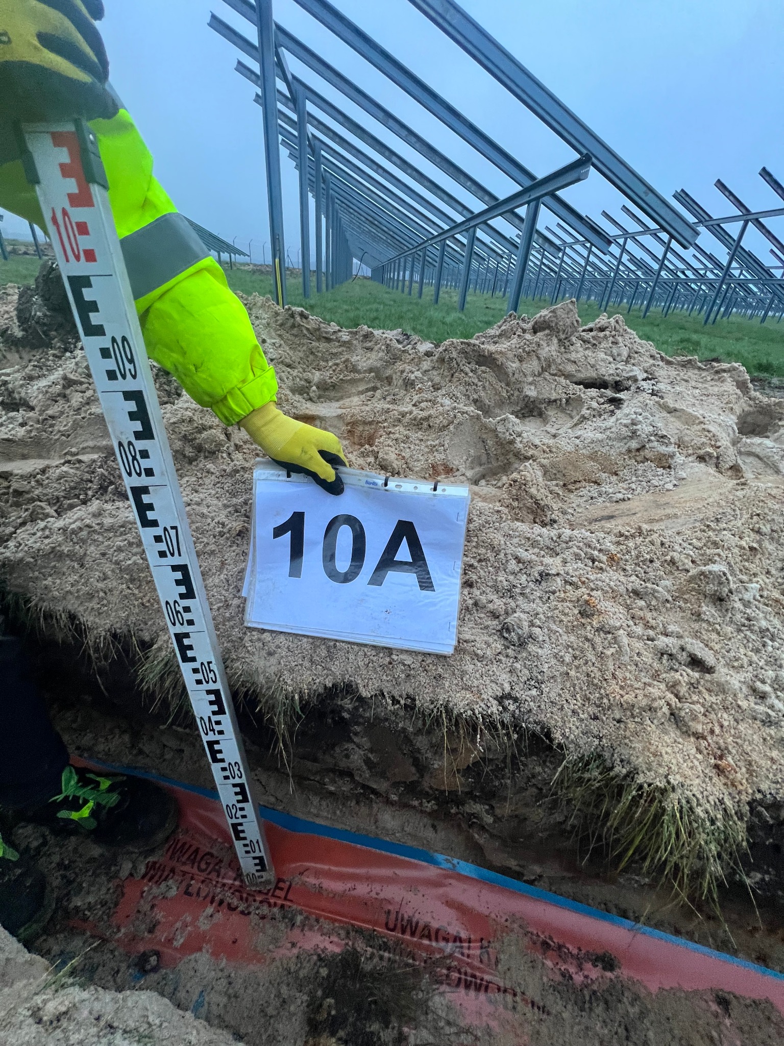 Pomiar głębokości wykopu pod instalację paneli fotowoltaicznych, widoczna łata miernicza, karta z oznaczeniem '10A' i zabezpieczenie kabla w wykopie, w tle konstrukcja wsporcza pod panele.