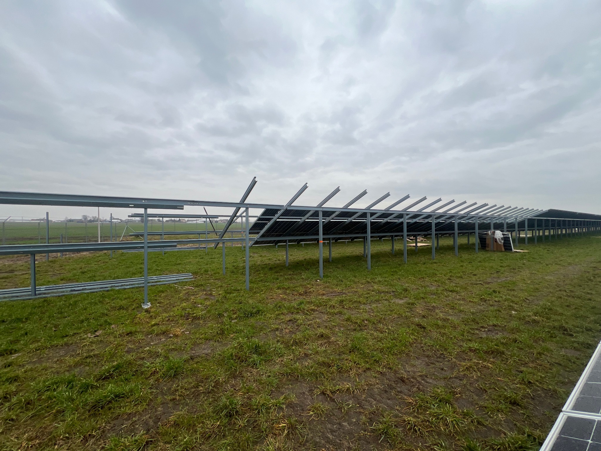 Stalowa konstrukcja wsporcza pod panele fotowoltaiczne na trawiastym terenie w pochmurny dzień, widoczne częściowo zamontowane panele i ogrodzenie w tle.