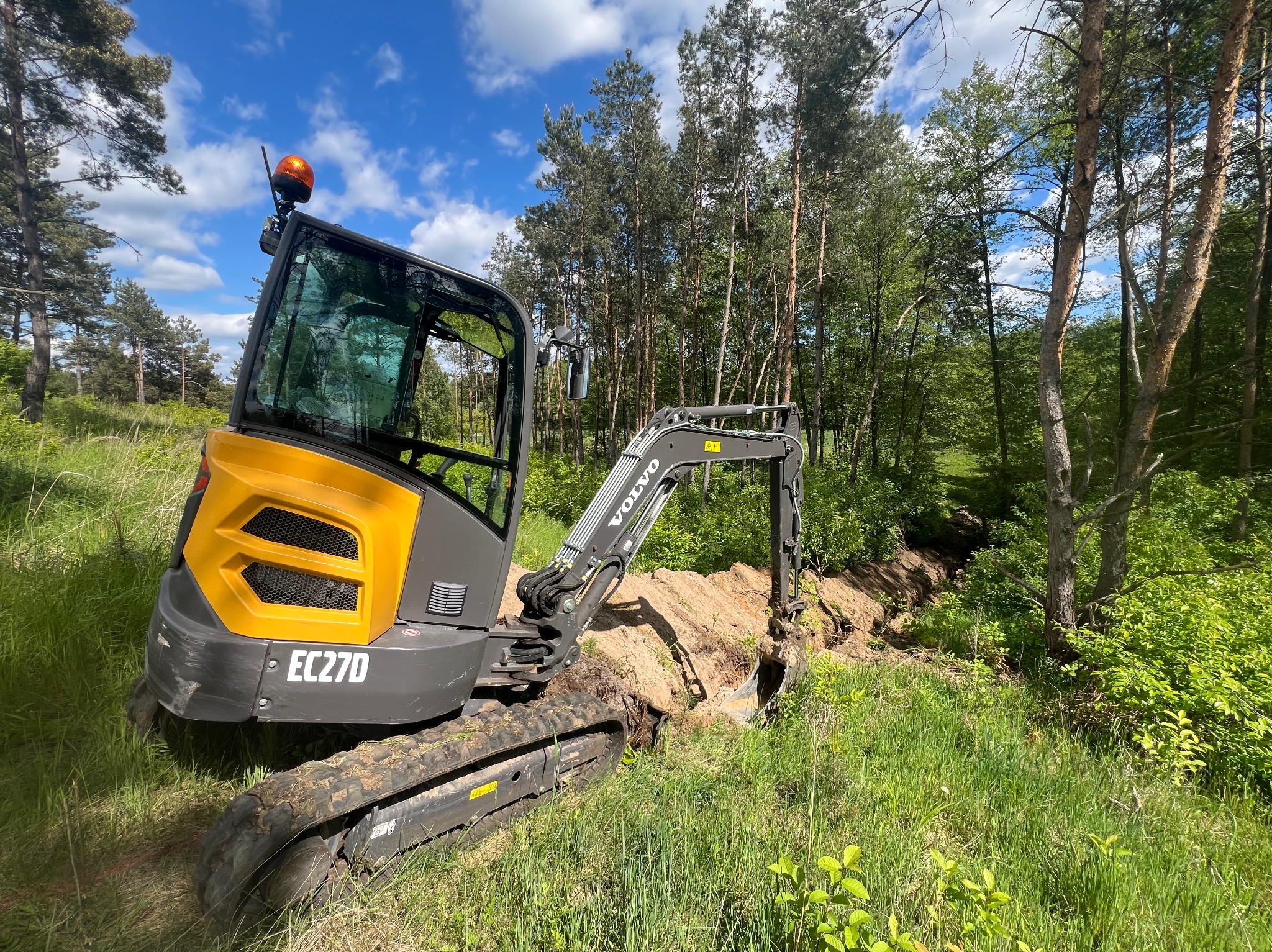 Żółto-szara minikoparka Volvo EC27D kopie rów na zalesionym terenie. Widoczne ślady błota na gąsienicach i świeżo wykopana ziemia. Błękitne niebo z chmurami w tle.