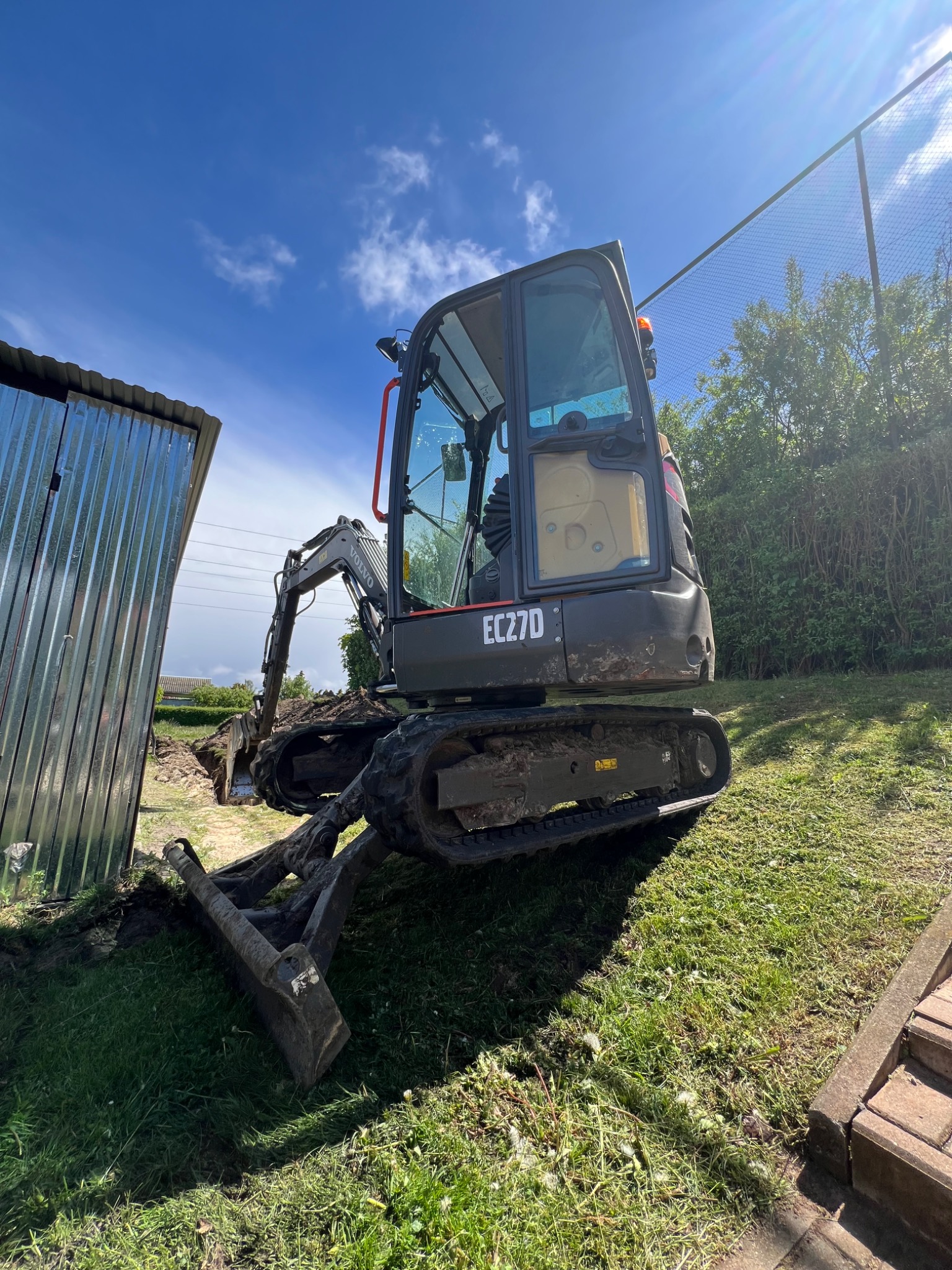 Koparka Volvo EC27D w trakcie prac ziemnych w Koninie. Widoczny wykop, fragment blaszanej konstrukcji i trawiasta skarpa pod błękitnym niebem.