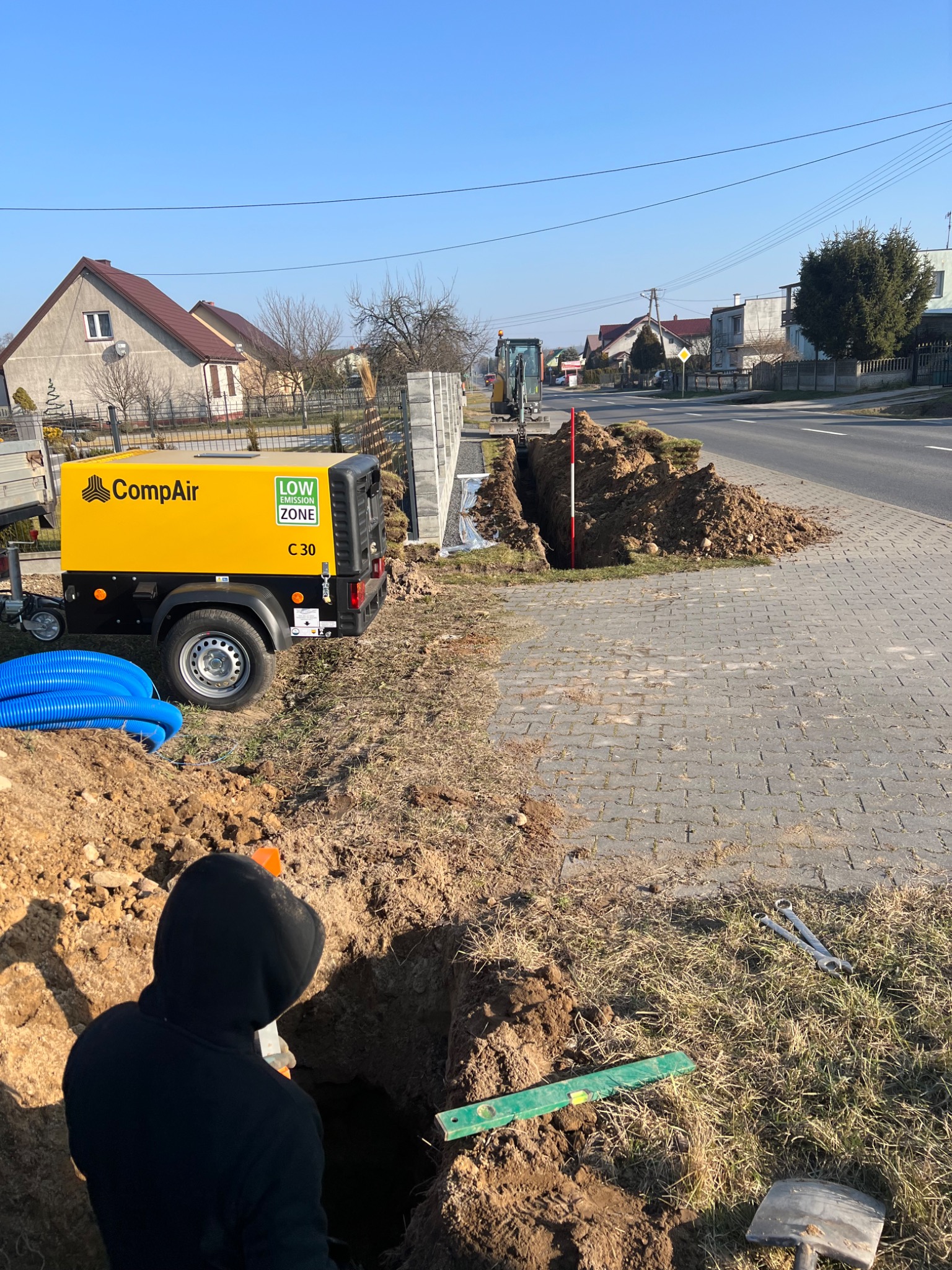 Wykop pod przyłącze, widoczna żółta sprężarka CompAir, niebieskie rury, poziomica, łopata i klucze. W tle domy i mini koparka na tle błękitnego nieba.