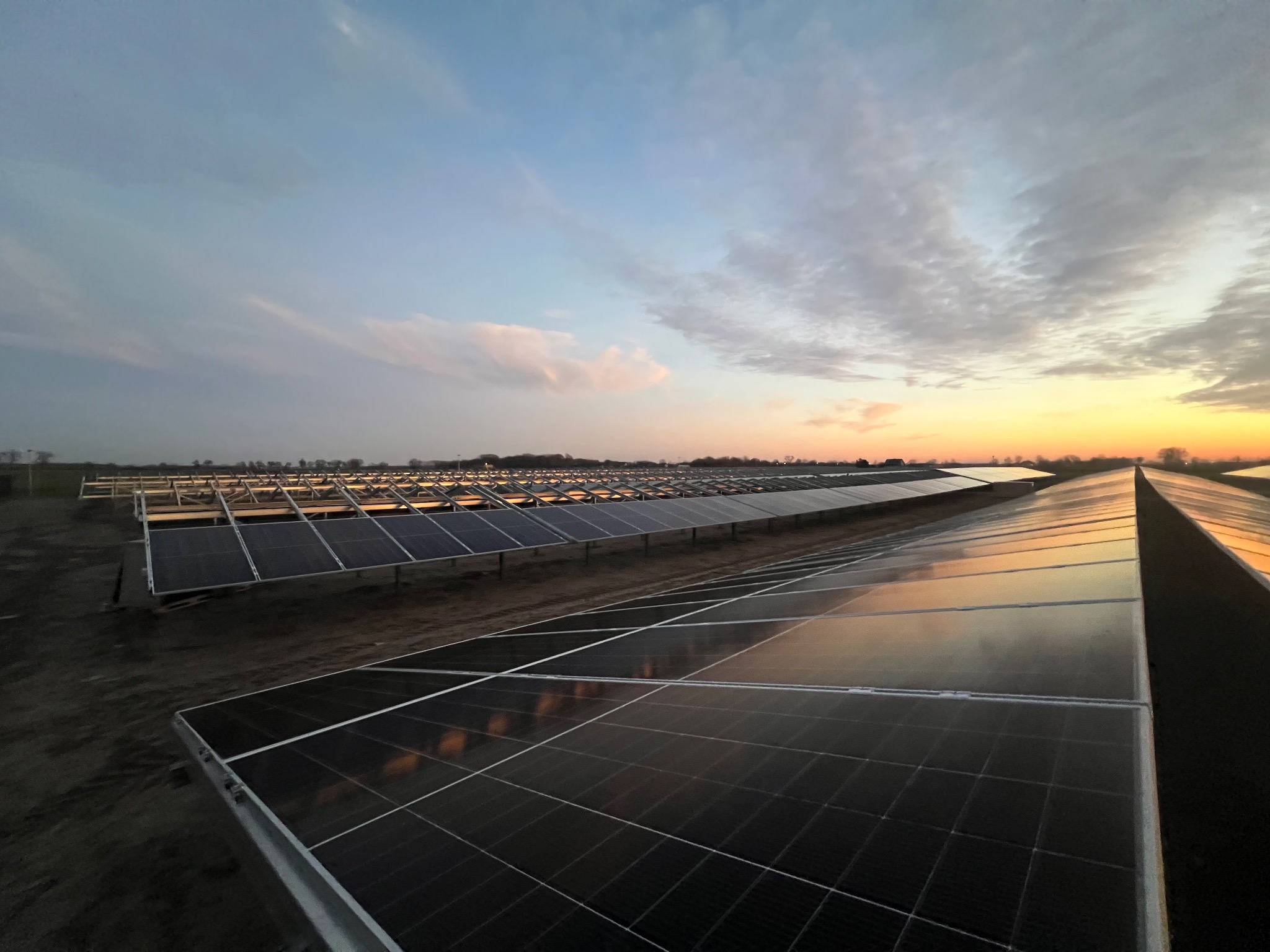Rozległa farma paneli słonecznych o zmierzchu, z widocznymi panelami odbijającymi niebo i zachodzące słońce. Ujęcie z niskiego kąta podkreśla rozległość instalacji.