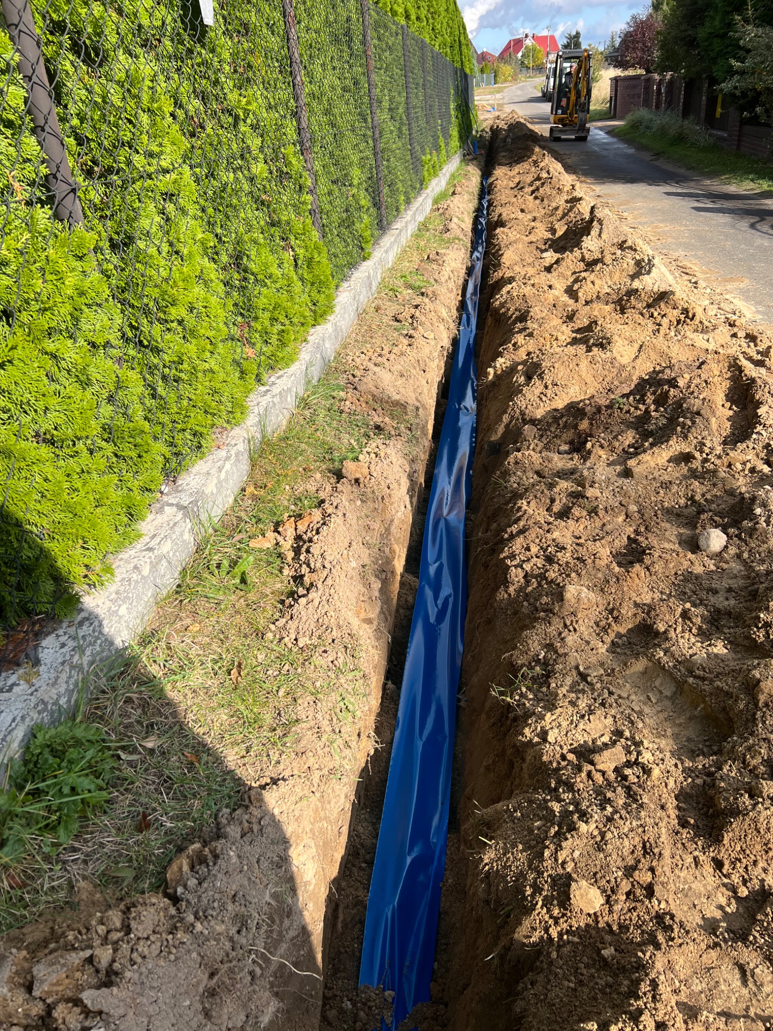 Wykop z zabezpieczeniem z niebieskiej folii, prawdopodobnie pod instalację sanitarną, z mini koparką w tle na drodze. Widok z góry, jasne światło słoneczne.