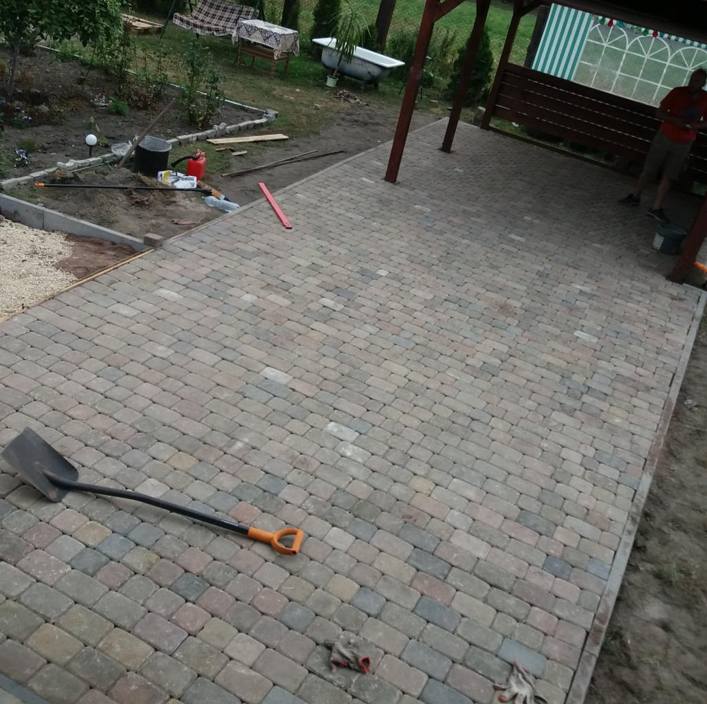 Ułożona kostka brukowa w odcieniach szarości i beżu pod zadaszeniem, widoczna łopata i rękawice robocze, w tle ogród z wanną.