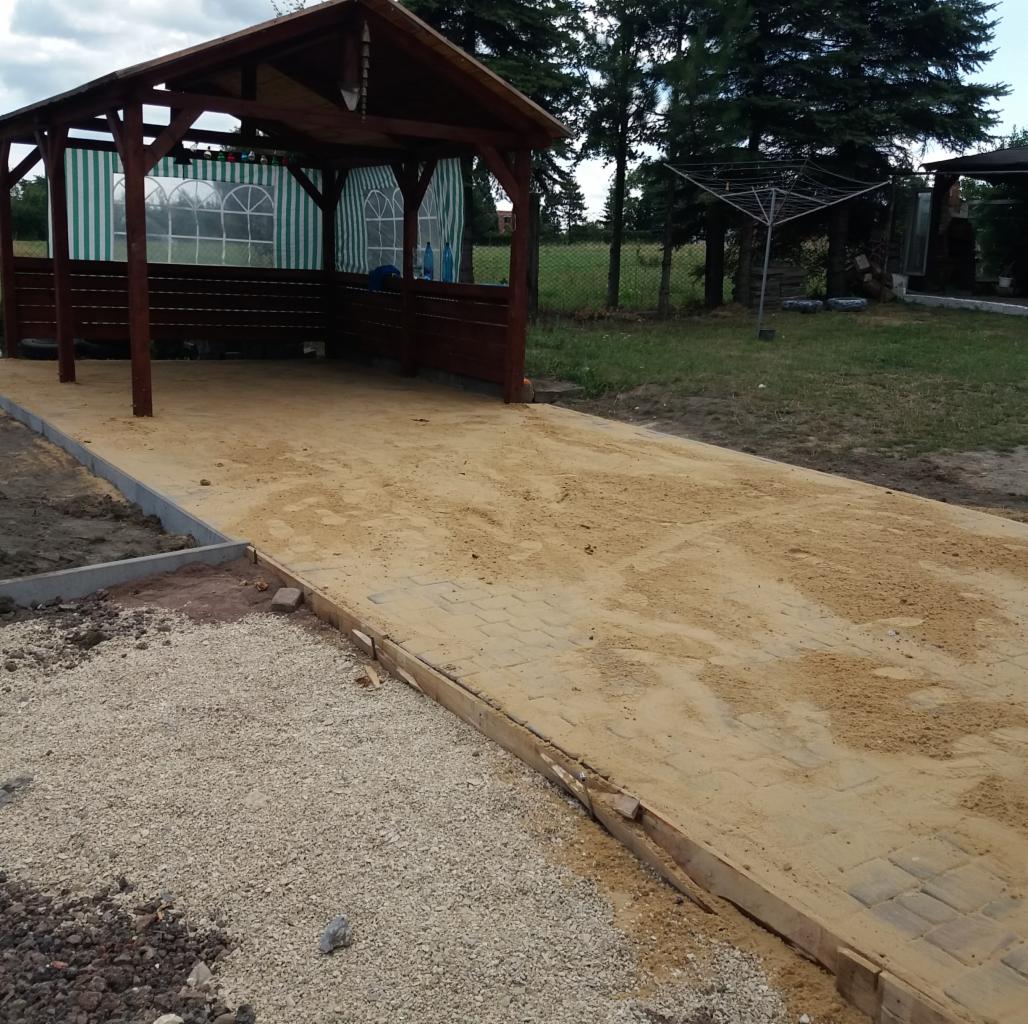 Układanie kostki brukowej pod drewnianą altaną z widocznymi fragmentami podsypki piaskowej i podsypki z kruszywa.