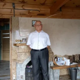 Mężczyzna w okularach, białej koszuli i ciemnych spodniach stoi w rustykalnym wnętrzu z drewnianym sufitem i kamiennym kominkiem.