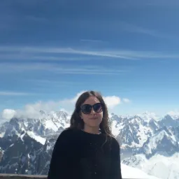 Portret kobiety w okularach na tle ośnieżonych gór i błękitnego nieba z chmurami. Ujęcie z lekkiej góry, podkreślające krajobraz.