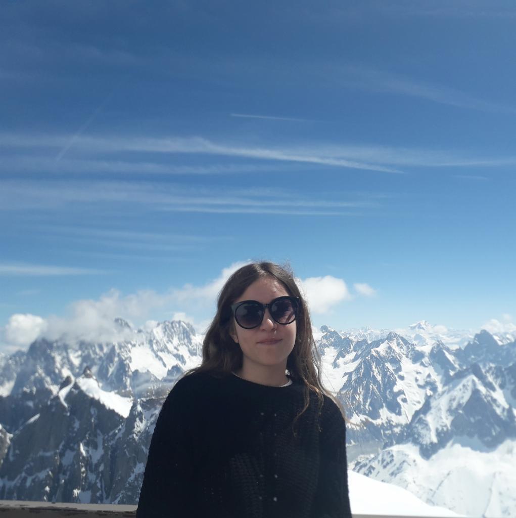 Portret kobiety w okularach na tle ośnieżonych gór i błękitnego nieba z chmurami. Ujęcie z lekkiej góry, podkreślające krajobraz.