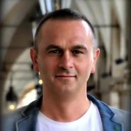 Portret mężczyzny w białej koszulce i dżinsowej marynarce na tle rozmytej architektury z łukami i oświetleniem.