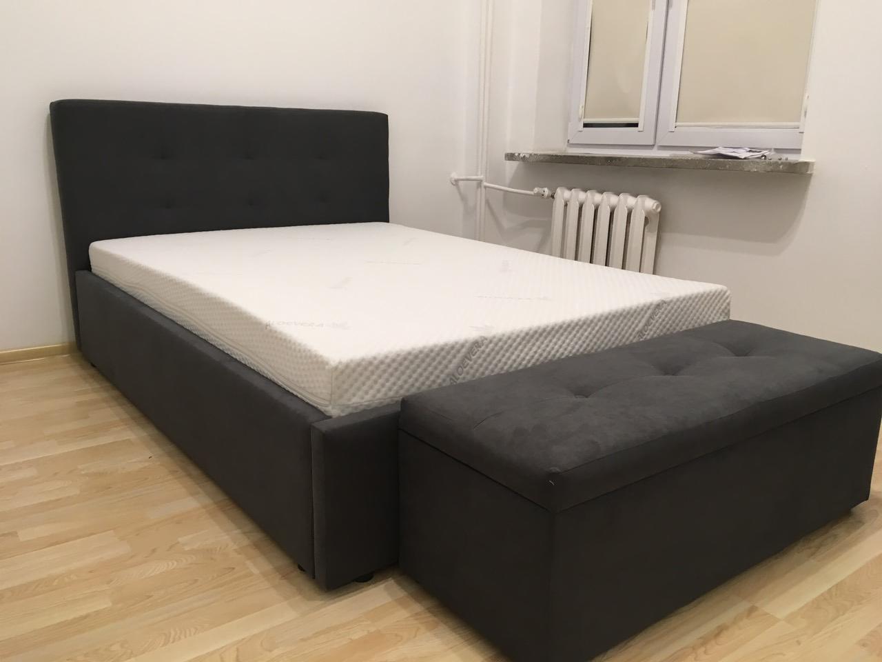 Szare tapicerowane łóżko z pojemnikiem, z materacem i tapicerowanym pufem w pokoju z parkietem i grzejnikiem pod oknem.