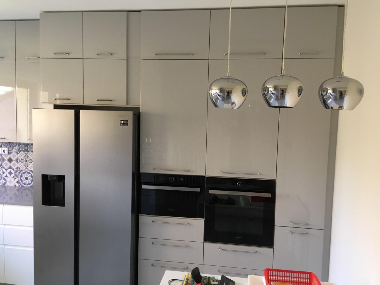 Jasnoszara kuchnia na wymiar z zabudowaną lodówką side-by-side i piekarnikami, oświetlona trzema chromowanymi lampami wiszącymi.