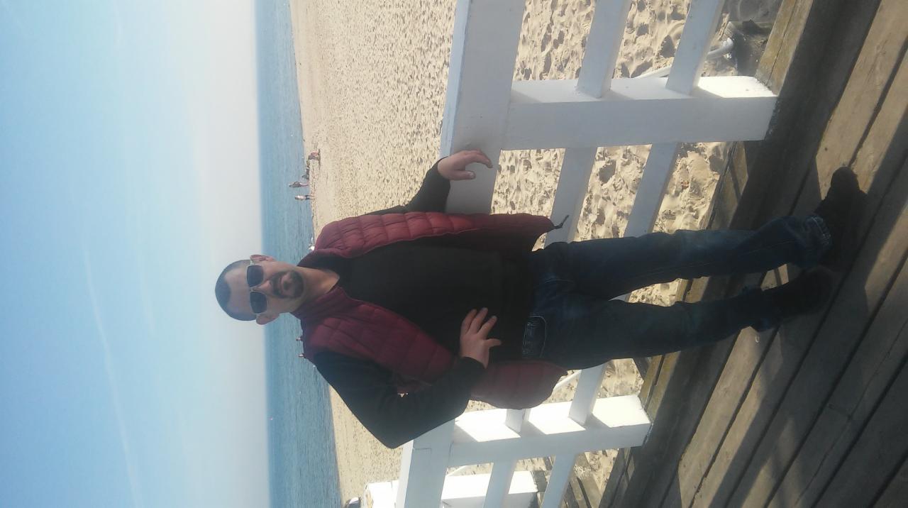 Mężczyzna w okularach przeciwsłonecznych opiera się o białą barierkę na drewnianym pomoście, z widokiem na plażę i morze w tle.