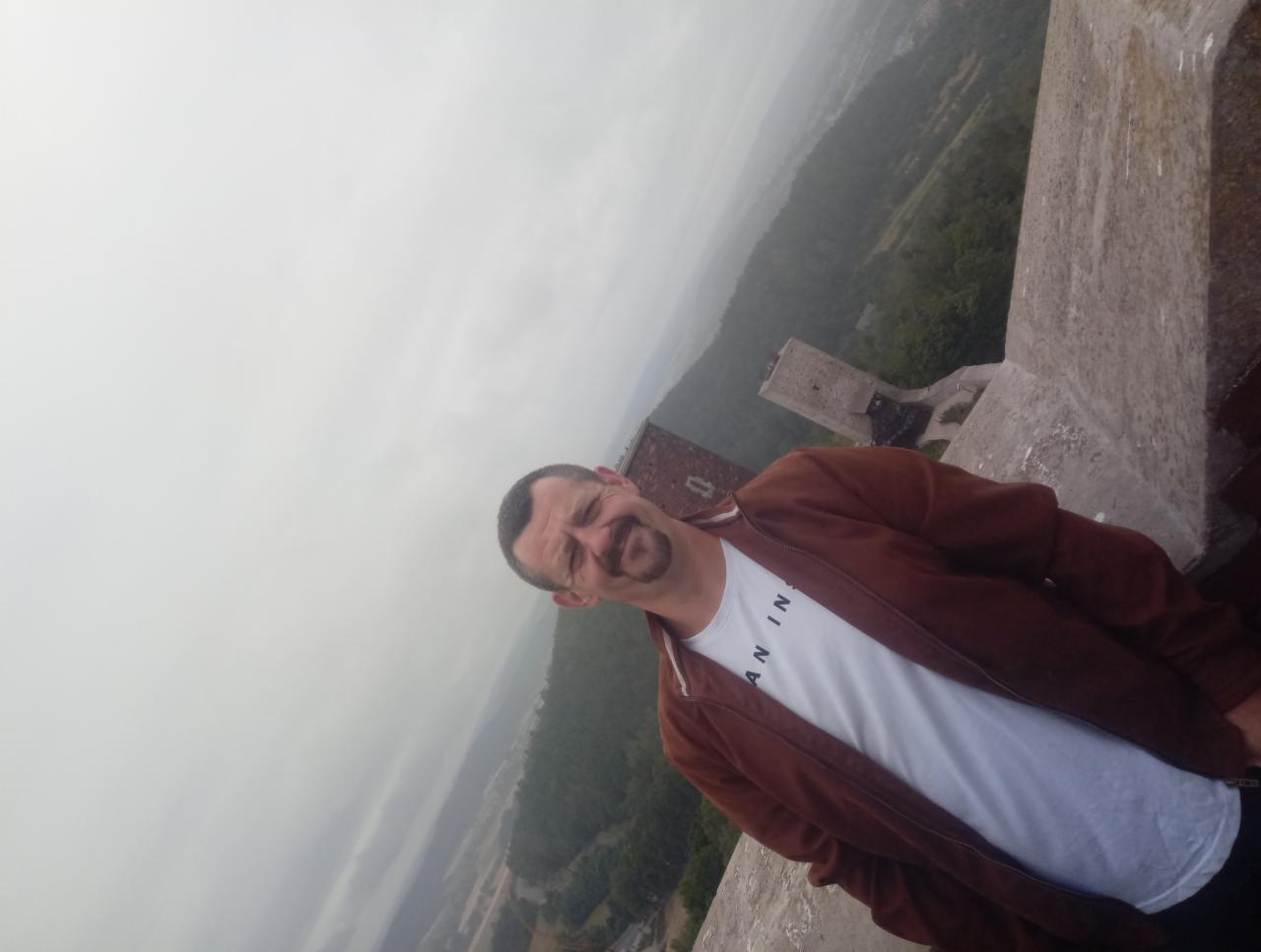 Mężczyzna w brązowej kurtce pozuje na tle zamku i mglistego krajobrazu. Nietypowa perspektywa z góry.