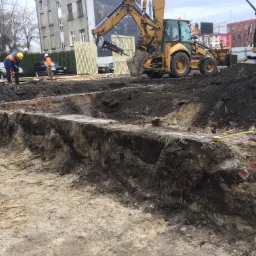 Wykop fundamentowy z widocznymi pracownikami w odblaskowych kamizelkach i żółtych kaskach, koparką CAT z młotem hydraulicznym i łyżką, oraz warstwami ziemi o różnej strukturze i kolorze. W tle...