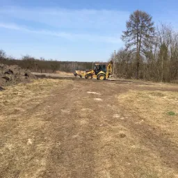 Żółta koparko-ładowarka na placu budowy z pryzmą ziemi w tle, podczas niwelacji terenu w słoneczny dzień.