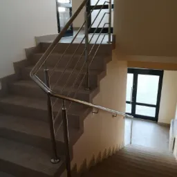 Wewnętrzna klatka schodowa z nowoczesną balustradą ze stali nierdzewnej, widok z góry na dół, z widocznymi drzwiami na obu poziomach.