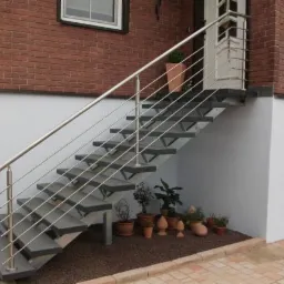 Zewnętrzne schody z szarymi stopniami i metalową balustradą z cienkich prętów, prowadzące do drzwi wejściowych, z roślinami w doniczkach u podstawy schodów.