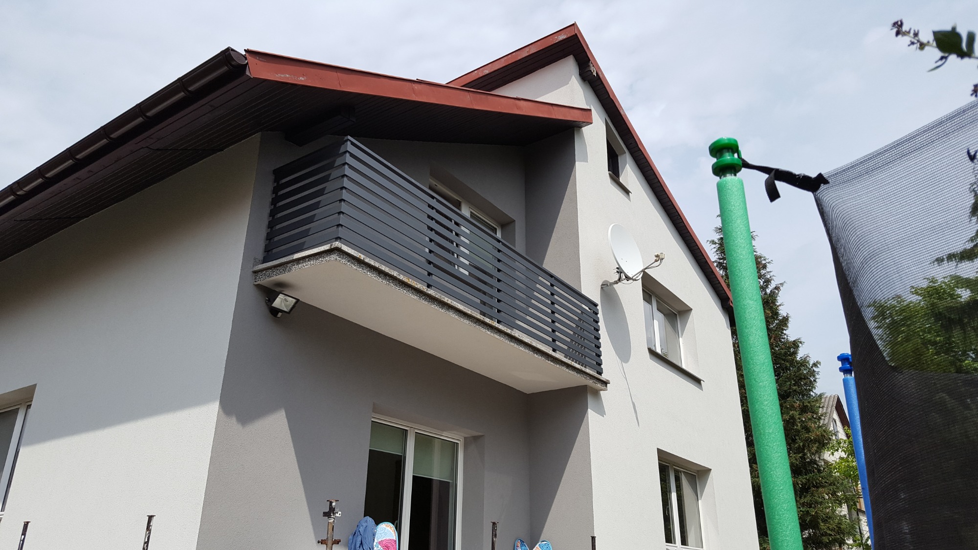 Szary dom z balkonem o czarnej, poziomej balustradzie z metalowych profili, widok z dołu, obok trampolina z zielonymi osłonami słupków.