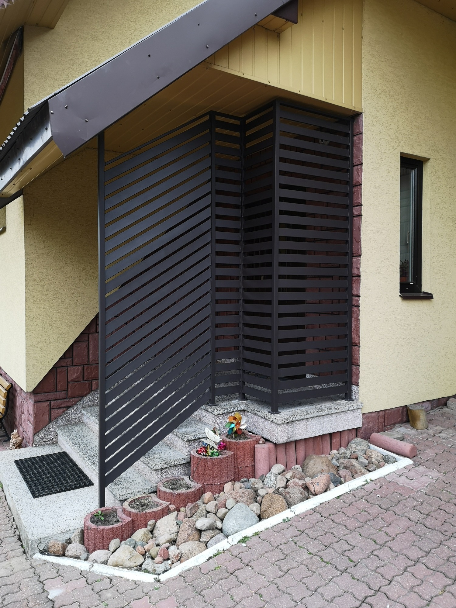 Nowoczesna, pozioma, metalowa osłona wejścia do domu z żółtą elewacją i kamienną podmurówką, z dekoracyjnymi kamieniami i donicami przed schodami.