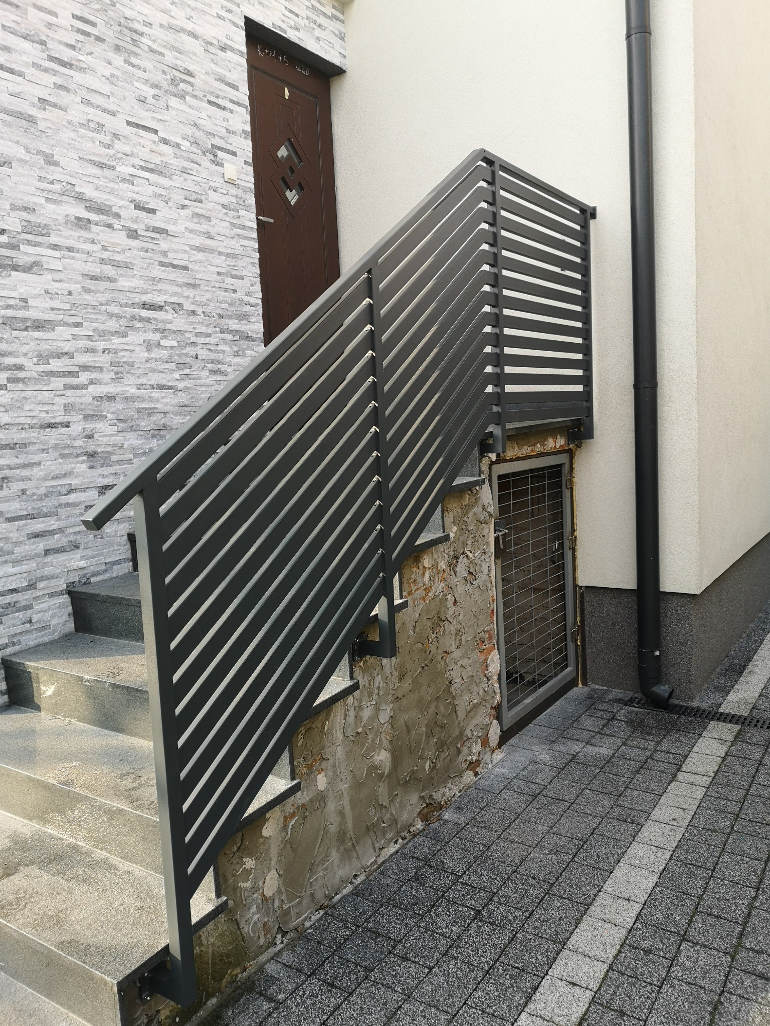 Nowoczesna, pozioma, metalowa balustrada schodowa w kolorze grafitowym, zamontowana przy wejściu do budynku z szarej cegły, z widocznymi schodami z szarego kamienia i kratą zabezpieczającą okienko...