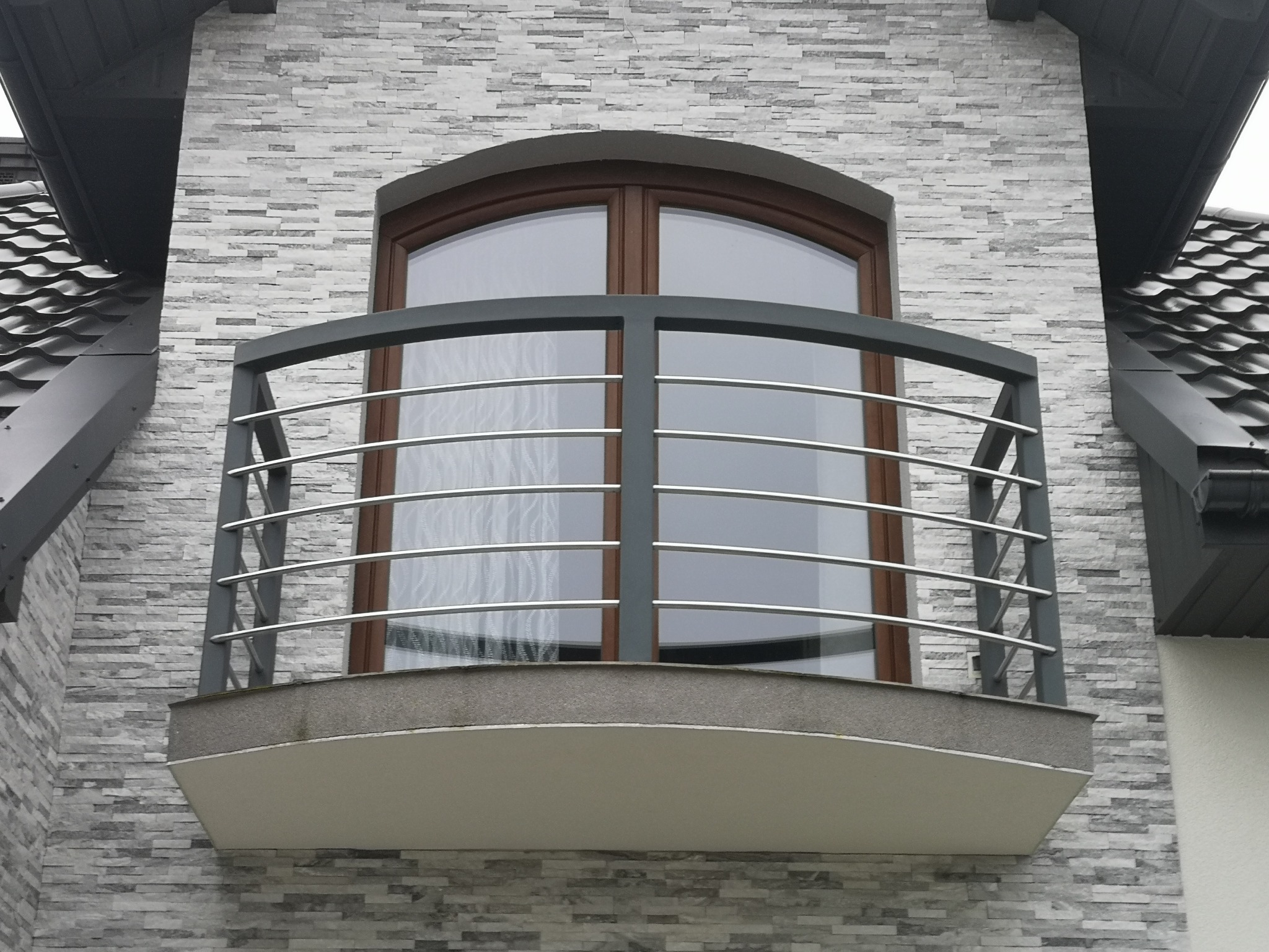 Nowoczesny balkon z szarymi, metalowymi barierkami i drewnianą ramą okienną na tle ściany z szarej cegły.