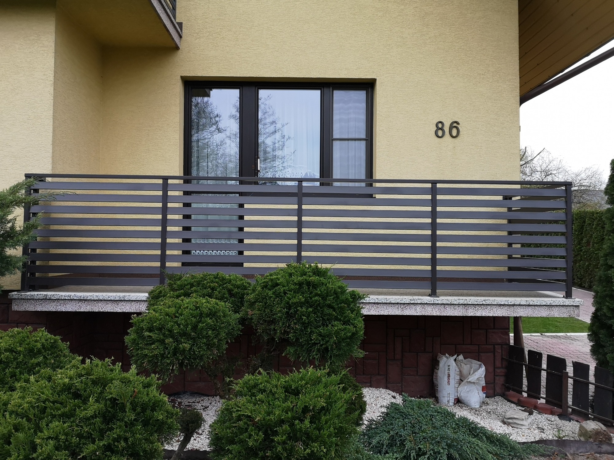 Nowoczesne ogrodzenie z poziomych, szarych profili metalowych przed budynkiem z numerem 86, otoczone zielonymi krzewami.