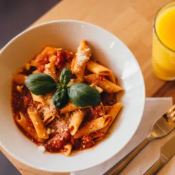 Penne w sosie pomidorowym z bazylią i parmezanem, podane w białej misce obok szklanki soku pomarańczowego oraz sztućców na drewnianym stole.