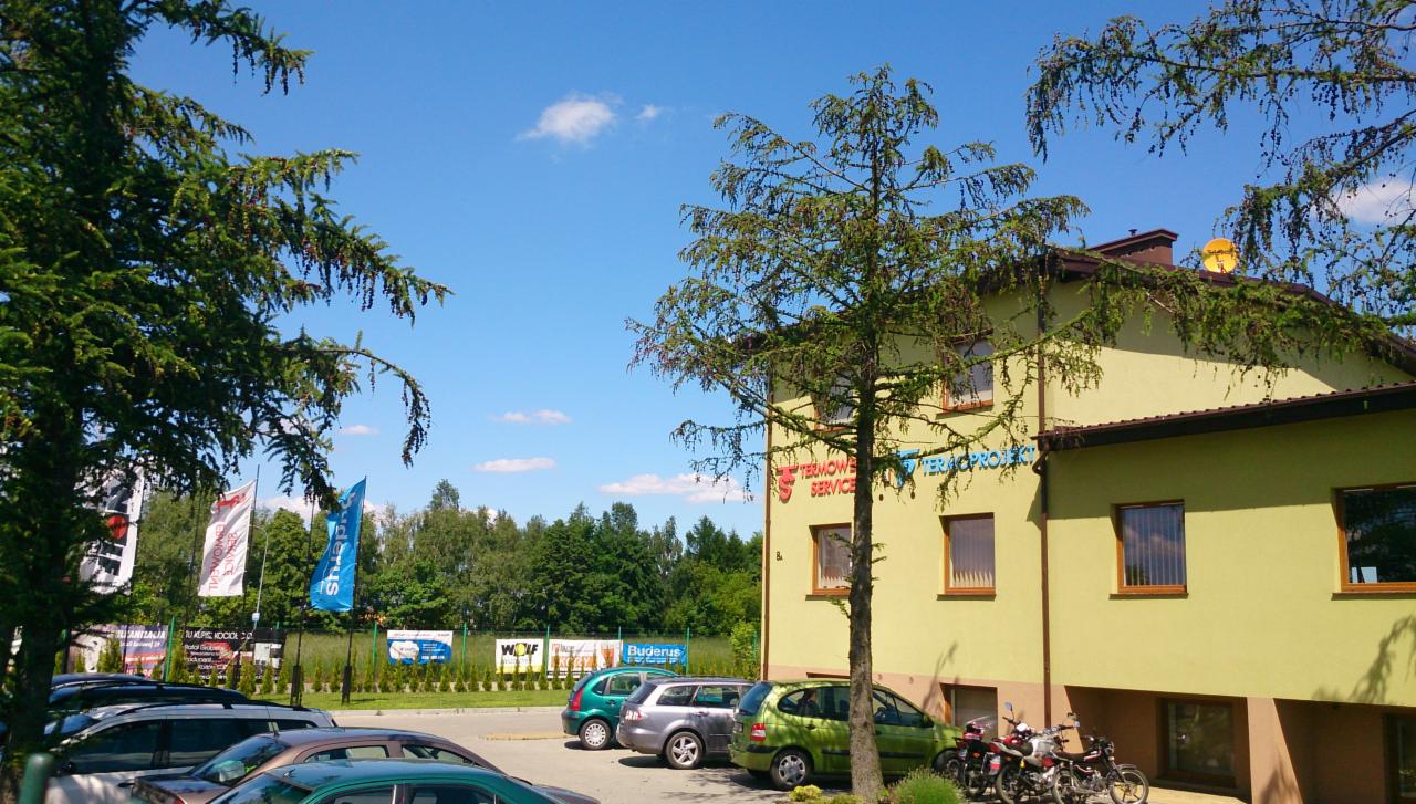Budynek firmy Termo-Project z logiem na elewacji, zaparkowane samochody i motocykle, widoczne banery reklamowe i drzewa w słoneczny dzień.