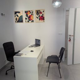 Poradnia Dietetyczna mgr Adrianna Barczyńska - Jasne biuro: białe biurko z laptopem, czarne krzesła, grafiki na ścianie. Minimalistyczny wystrój, lampa sufitowa. Przestrzeń gabinetu.