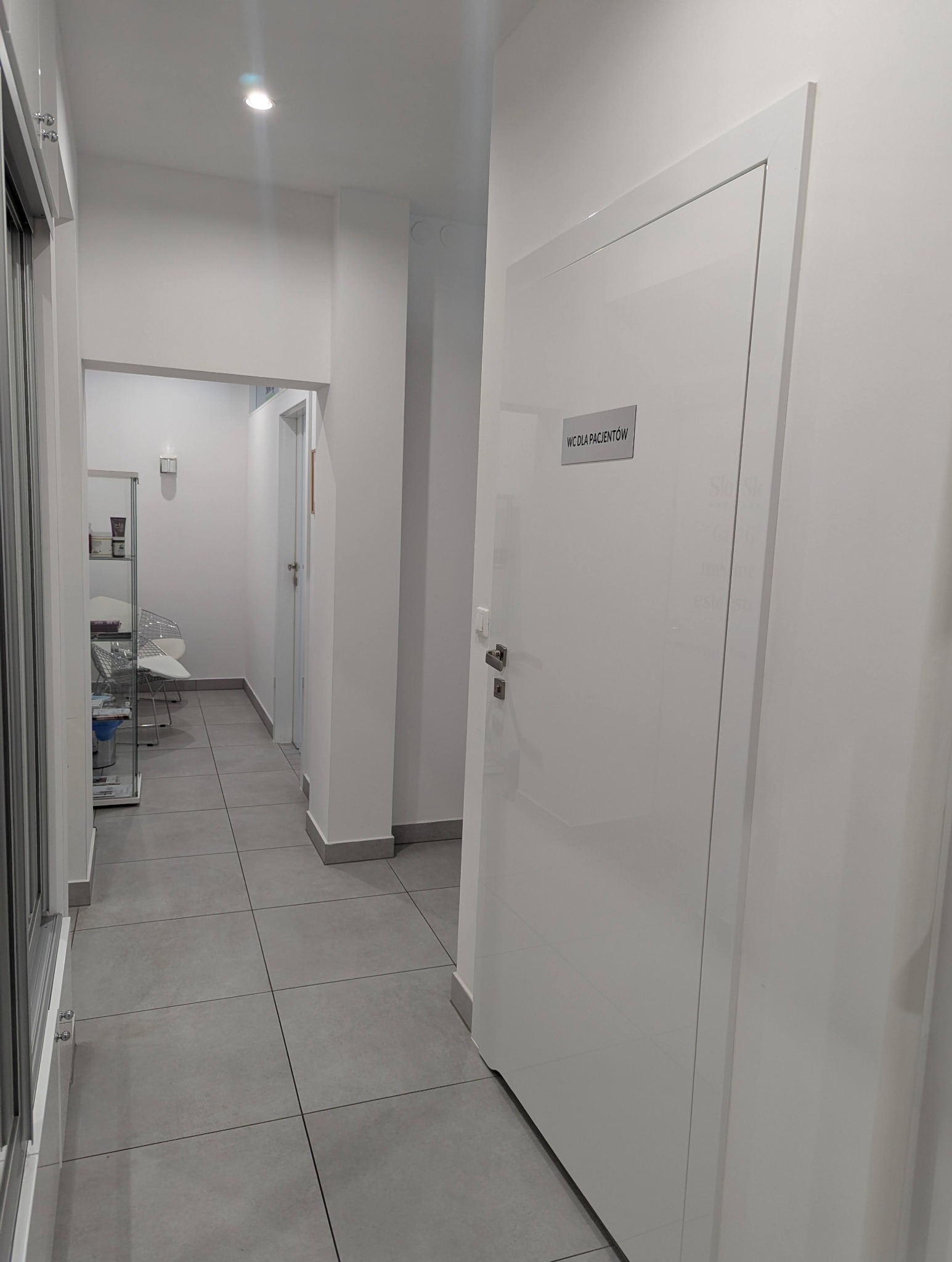 Wnętrze gabinetu z białymi ścianami, szarą podłogą i drzwiami z napisem 'WC dla Pacjentów'. Widok na korytarz z recepcją w tle. Minimalistyczny design.