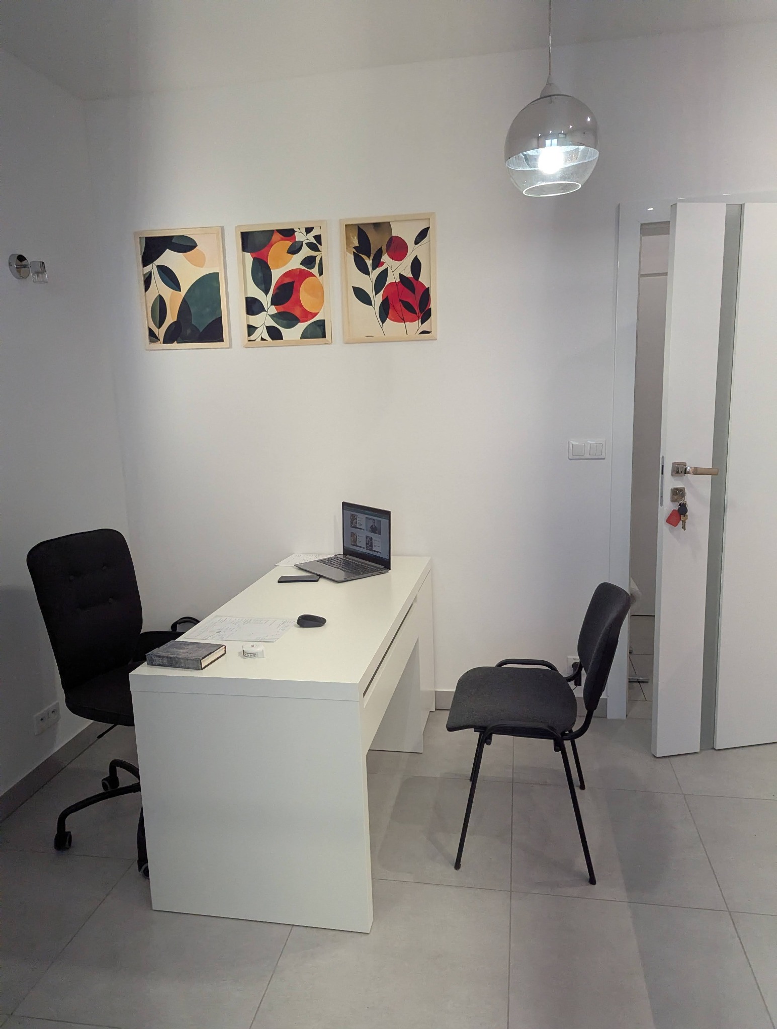 Jasne biuro: białe biurko z laptopem, czarne krzesła, grafiki na ścianie. Minimalistyczny wystrój, lampa sufitowa. Przestrzeń gabinetu.