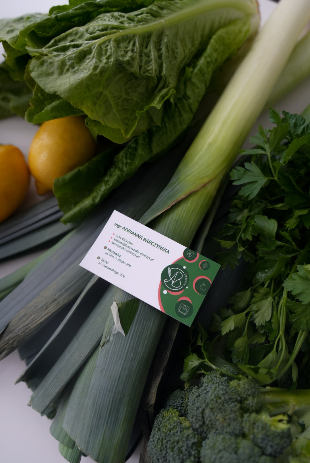 Kompozycja świeżych warzyw (sałata, pory, brokuły, natka pietruszki) i cytryn z wizytówką mgr Adrianny Barczyńskiej, dietetyka, na białym tle.