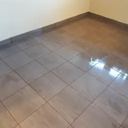 Wykończona podłoga z szarych, kwadratowych płytek ceramicznych, z ciemnoszarą listwą przypodłogową przy jasnej ścianie.