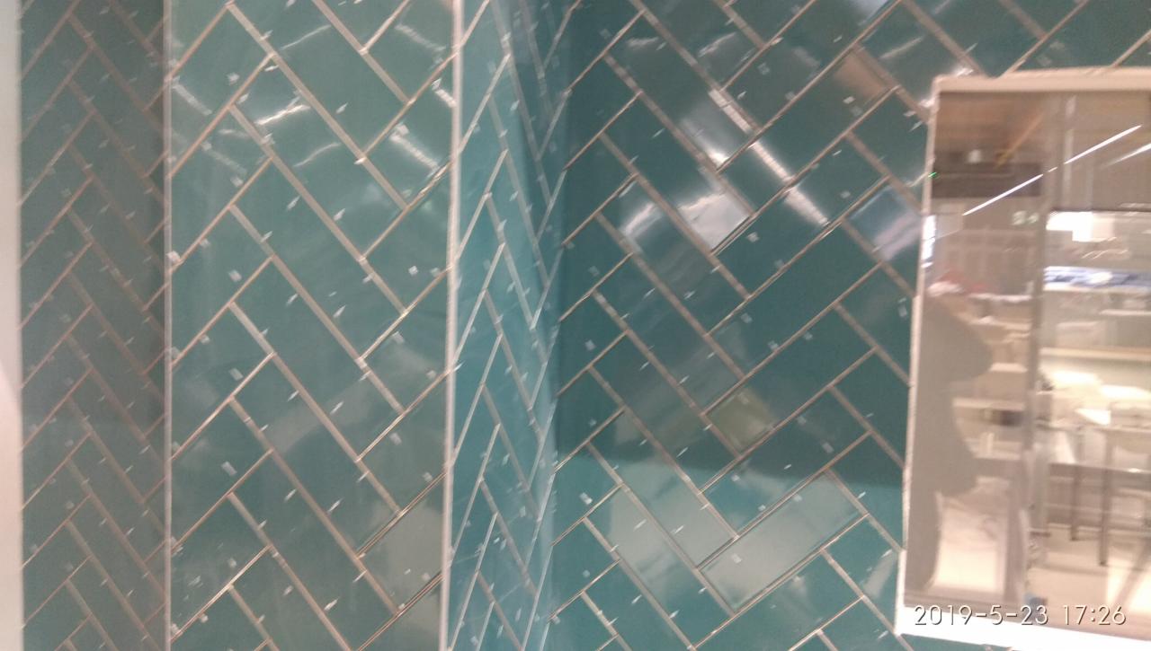 Ściana wyłożona błyszczącymi, turkusowymi płytkami ułożonymi we wzór jodełki, widoczny narożnik i fragment framugi drzwi